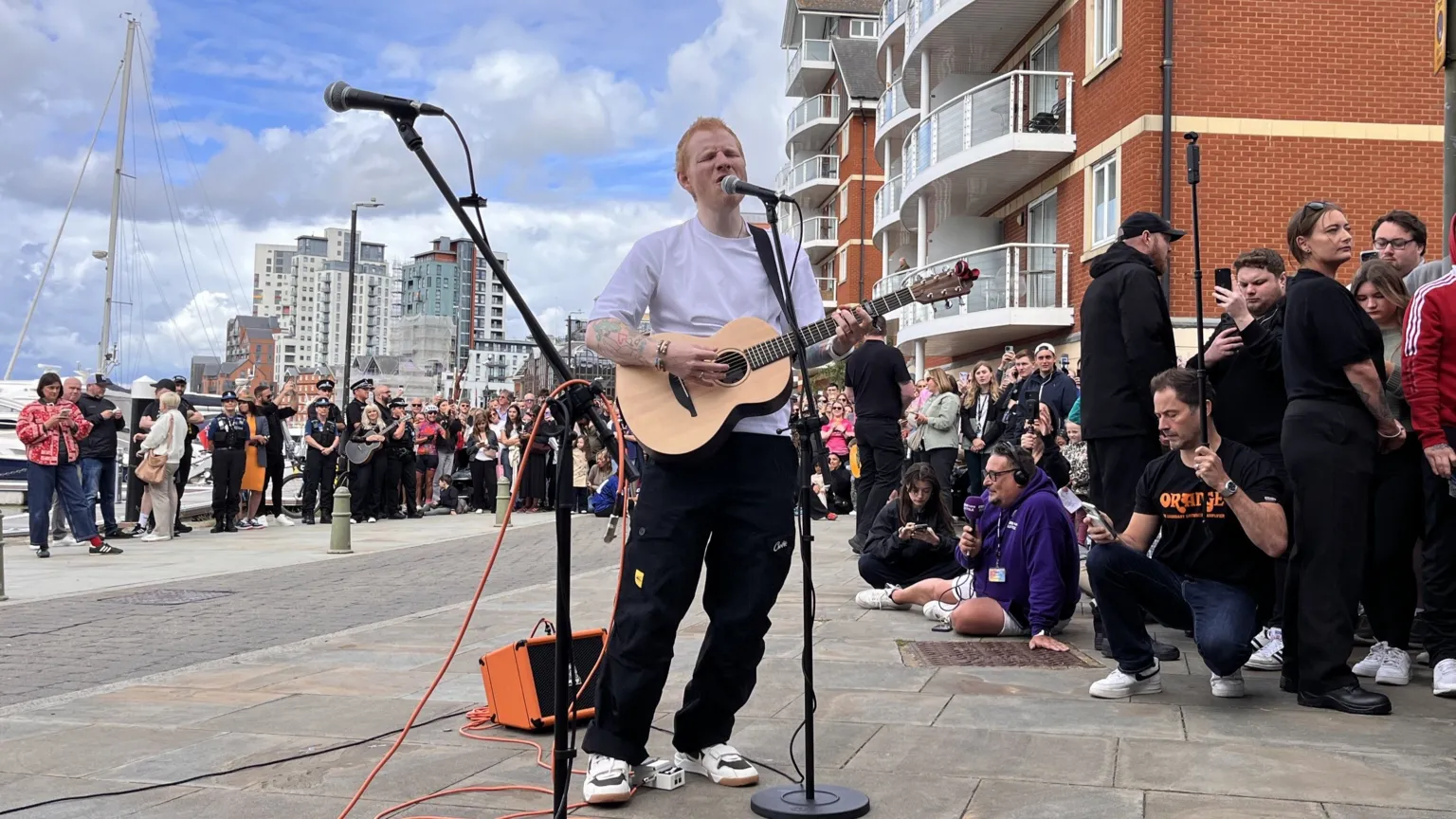 Shaun Whitmore/BBC A man with shaved red hair wearing a white T-shirt plays an acoustic guitar and sings into a microphone. There is a crowd of people around him. Some buildings are in the distance as well as boats on a marina.