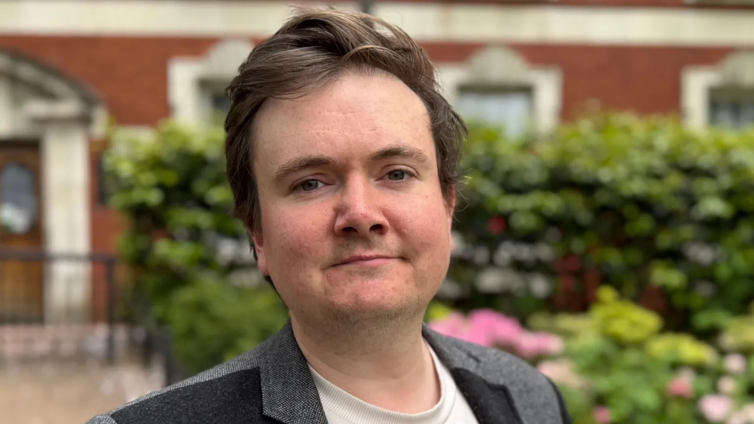 Liberal Democrats Jake Austin smiles for the camera in a large garden with a public building behind him. He has short dark hair and wears a dark blazer over a white top.