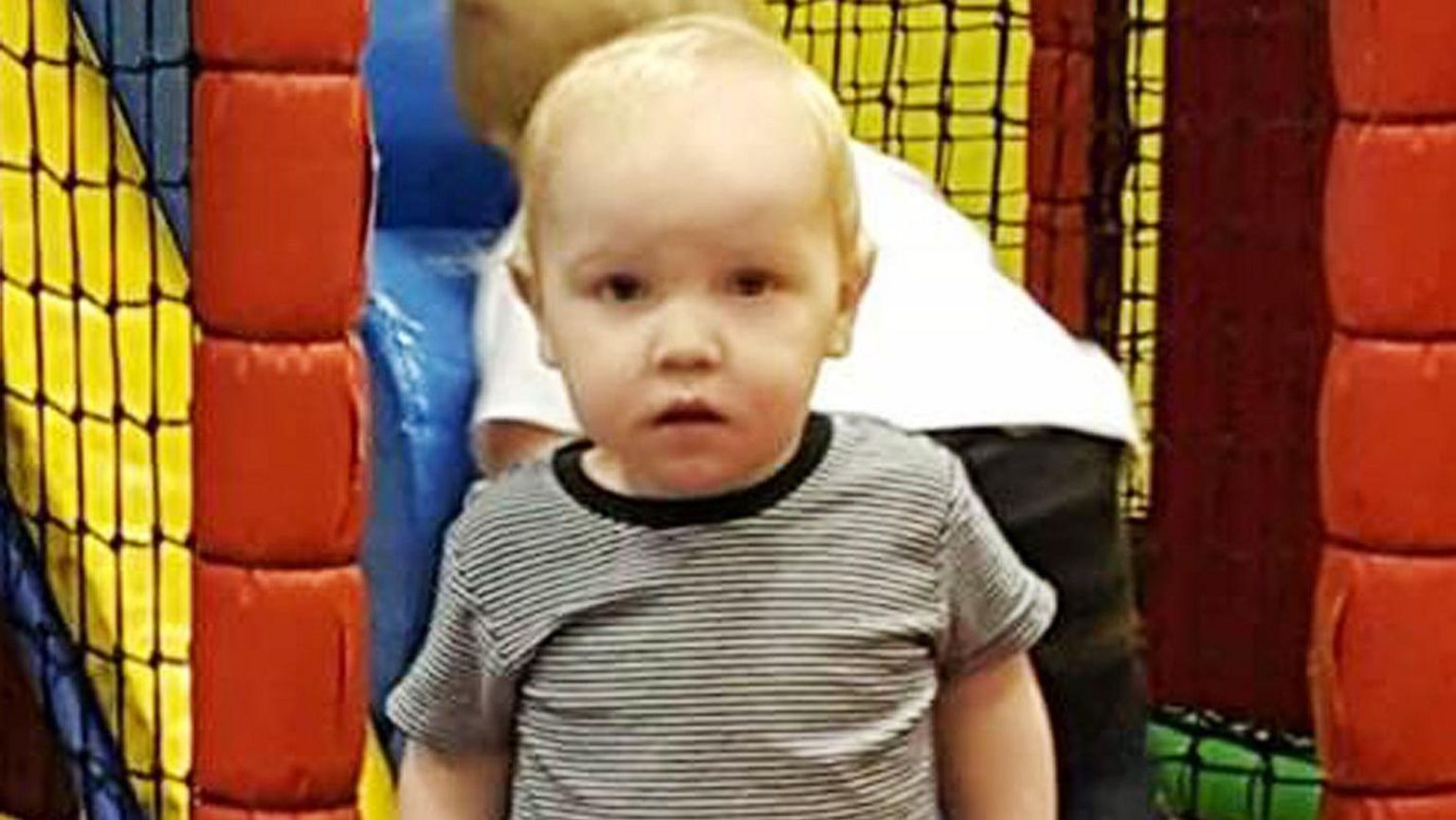 Toddler Harry Studley has short blonde hair and is wearing a black and white striped top. He is standing in the middle of a soft play centre.