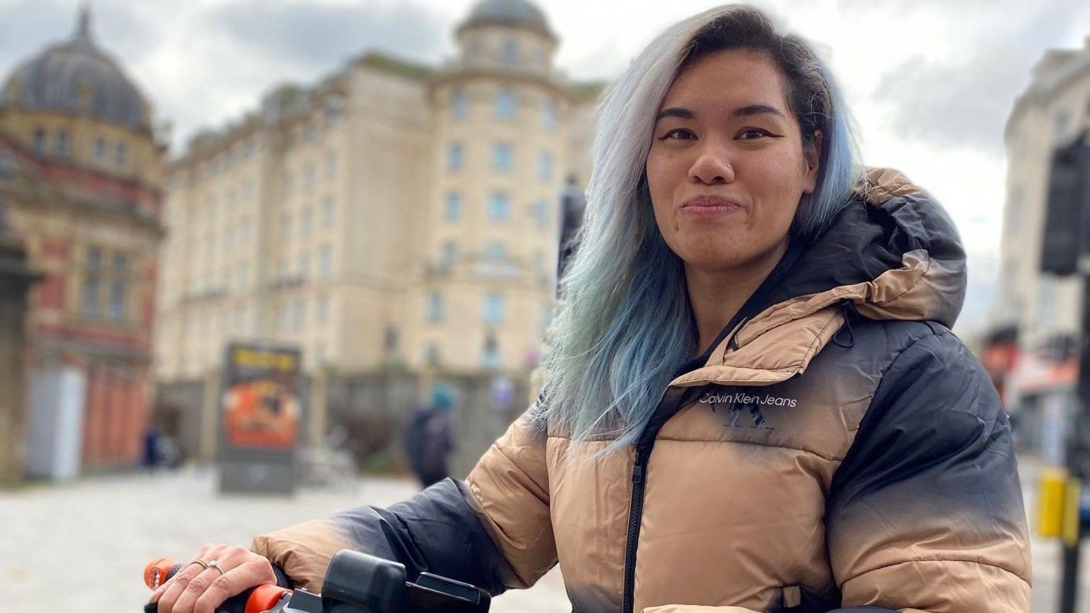 Jess wearing a cream and dark blue puffer jacket with blue hair standing with an e-scooter smiling at the camera