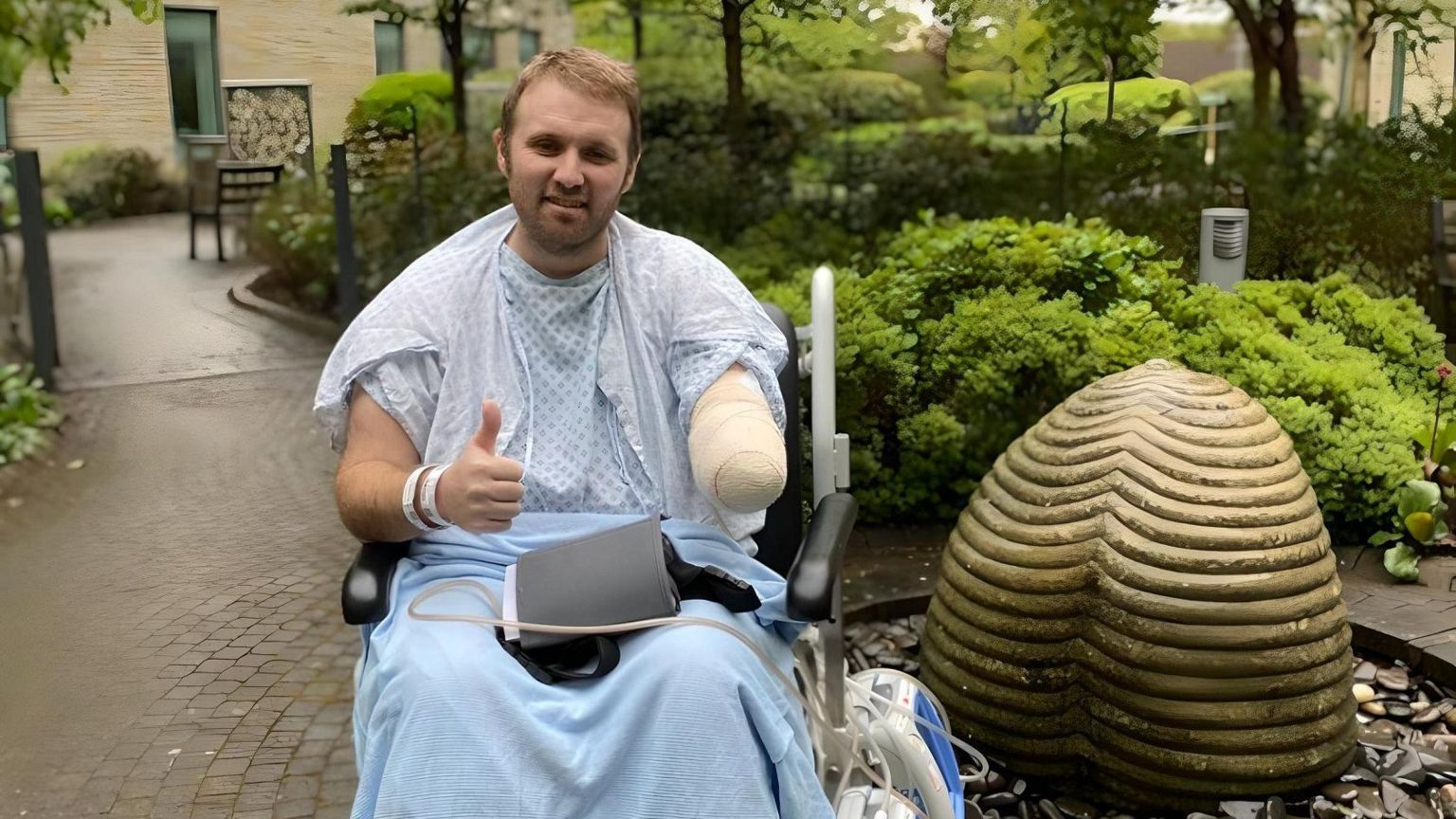 John is pictured outside in a wheelchair, wearing a hospital gown with a blue blanket over his legs. He is smiling at the camera and giving a thumbs up. 