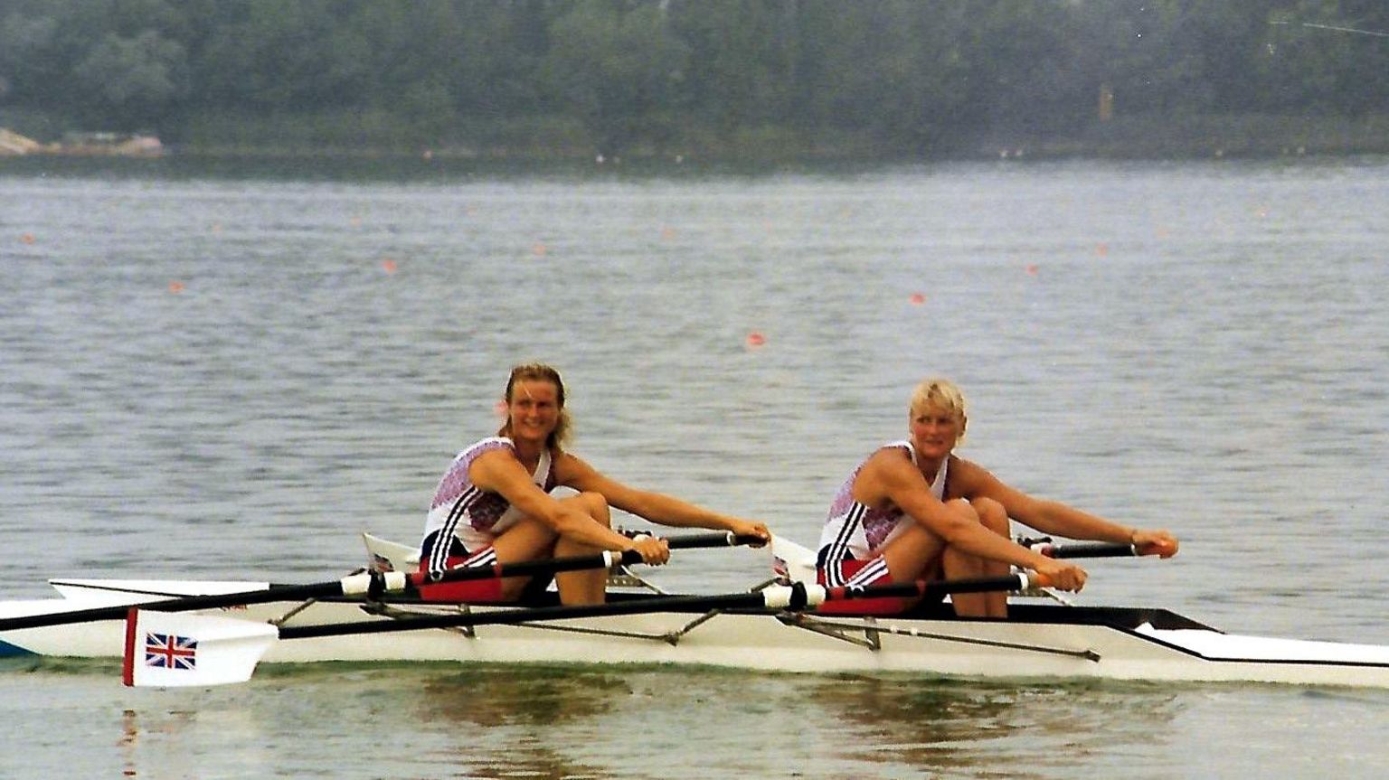 Annabel Eyres rowed and Alison Gill in a rowing boat on the water at the Barcelona Olympics. They are wearing red white and blue outfits and have British flags on their oars