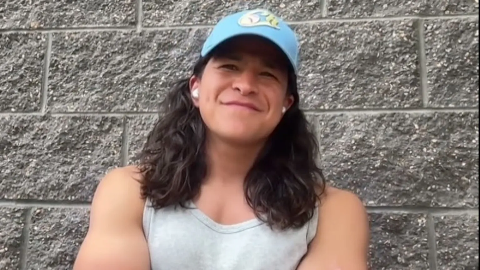Cristo Fernandez wearing a blue cap and grey singlet leaning against a stone wall.