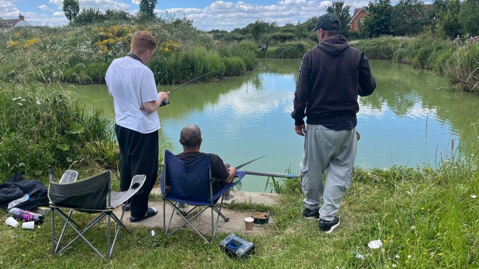 Three men fishing together at a lake, with their backs to the camera as they look out across the water. Two are standing and the one in the middle is sitting in a camping chair