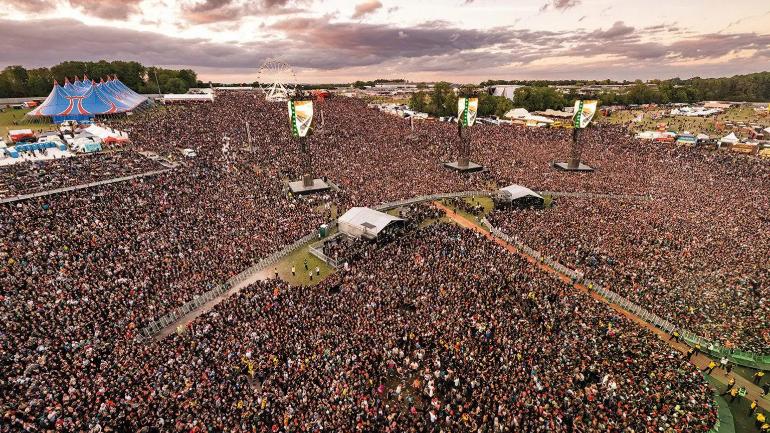 Sarah Louise Bennett/Download Festival An aerial shot of thousands of people at a festival, with a large blue performance tent in the top left and tall sound speakers in the crowd.