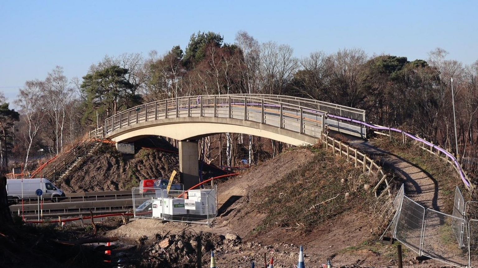 A building site featuring a central curved footbridge over a major road with woodland in the background