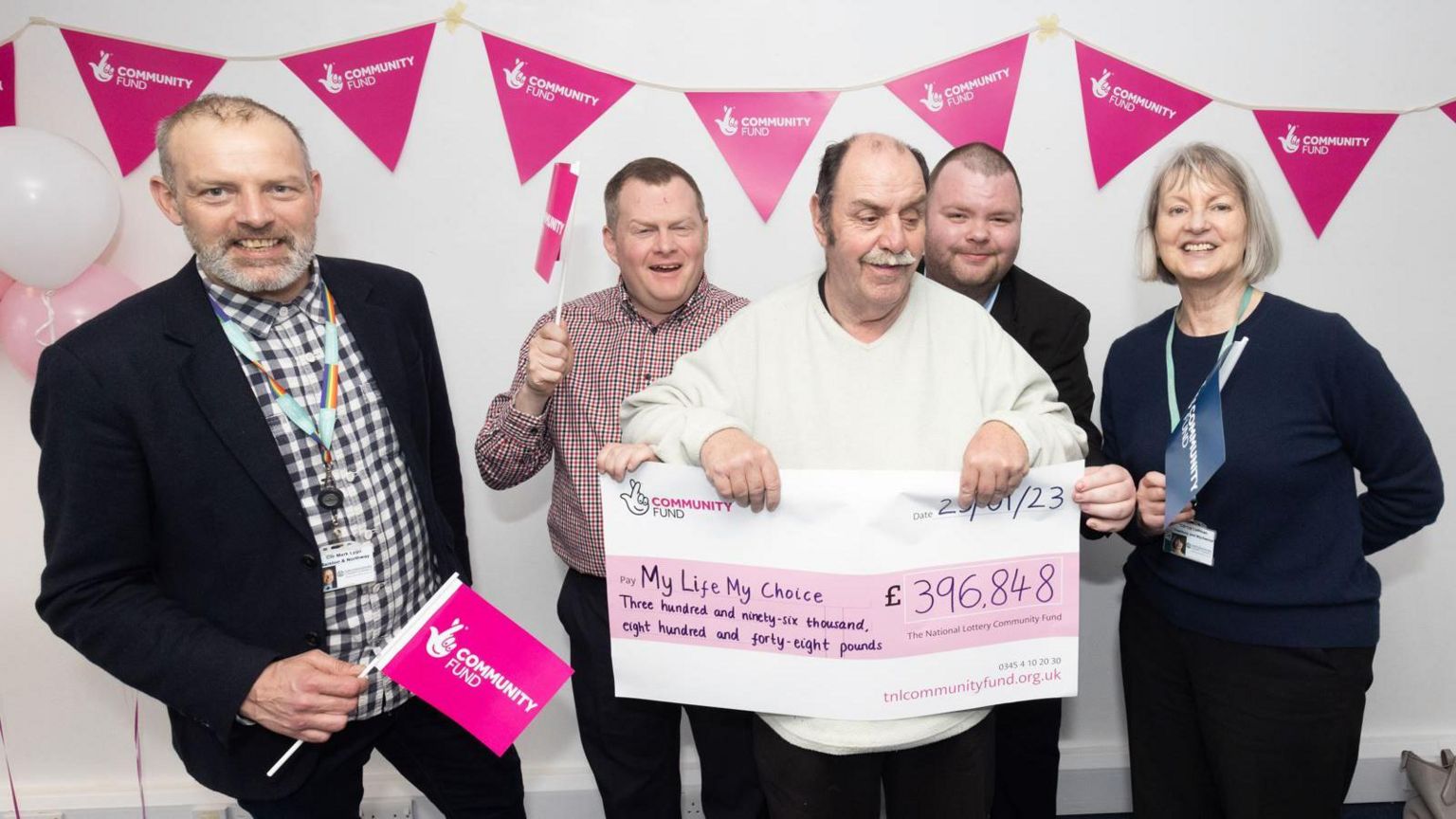 Michael Edwards at a Gig Buddy funding event in March 2023. He is in the middle of the frame, holding a check for £396.848. He wears a white long-sleeve top and dark trousers. On his left side is vice chair of Oxfordshire County Council Mark Lygo. He is wearing a black blazer and trousers and a shirt with blue and white squares. He is smiling at the camera and holding a pink flag with the name and logo of the Community Fund. On Michael’s right side is the council leader Liz Leffman, who is wearing dark long-sleeved top and trousers. She is also smiling at the camera and holding a blue flag which also appears to say Community Fund. There are two men behind Michael, one of which is waving a pink flag, the other one – smiling at the camera. The wall behind them is decorated with  Community Fund bunting. There are a pink and white balloon on the left corner.