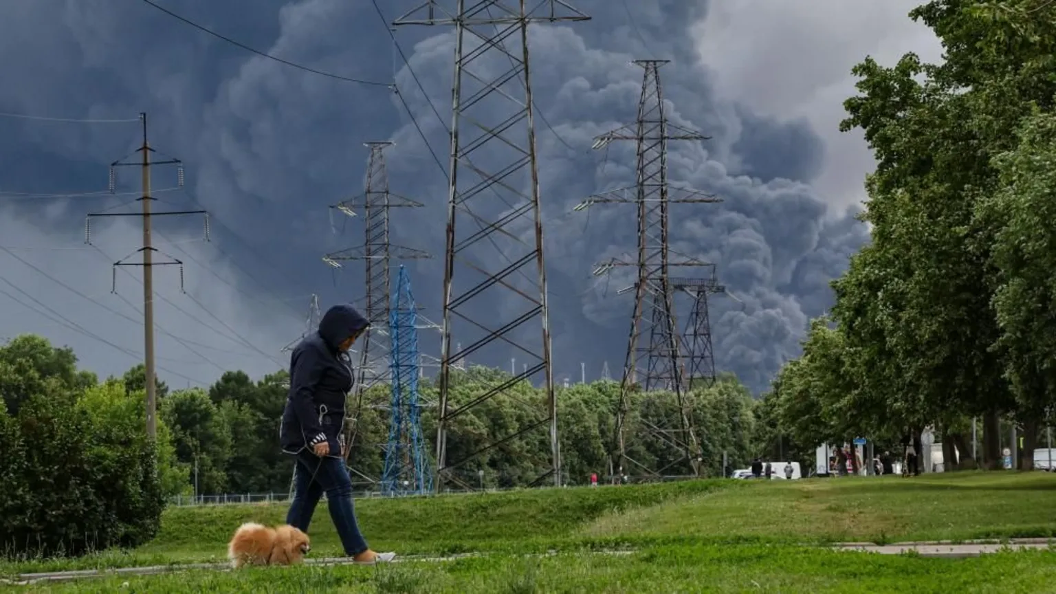 A photograph showing a person wearing a dark blue hooded jacket and jeans, walking a small, fluffy orange dog on a leash along a green, grassy path. In the midground, several tall metal electrical transmission towers stand in front of a line of green trees. Looming behind the trees and dominating the background sky is a massive, thick plume of dark grey smoke billowing from a large, unseen fire or explosion.