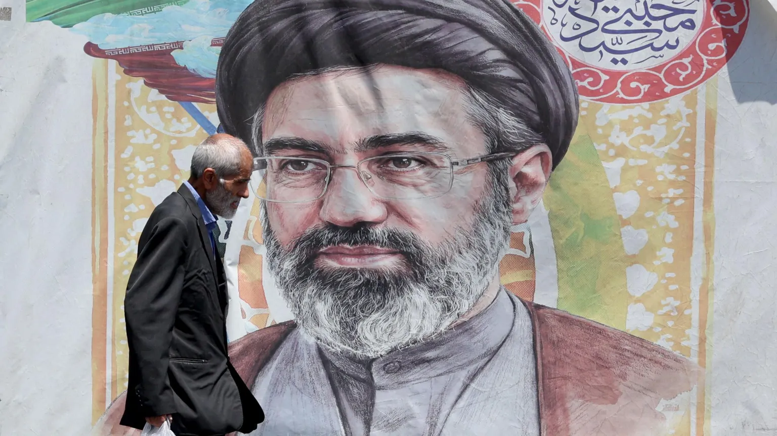 EPA A man with grey hair wearing a dark suit and blue shirt carrying a plastic bag walks past a poster of Supreme Leader Mojtaba Khamenei who has a grey beard and is wearing a grey buttoned shirt, mar