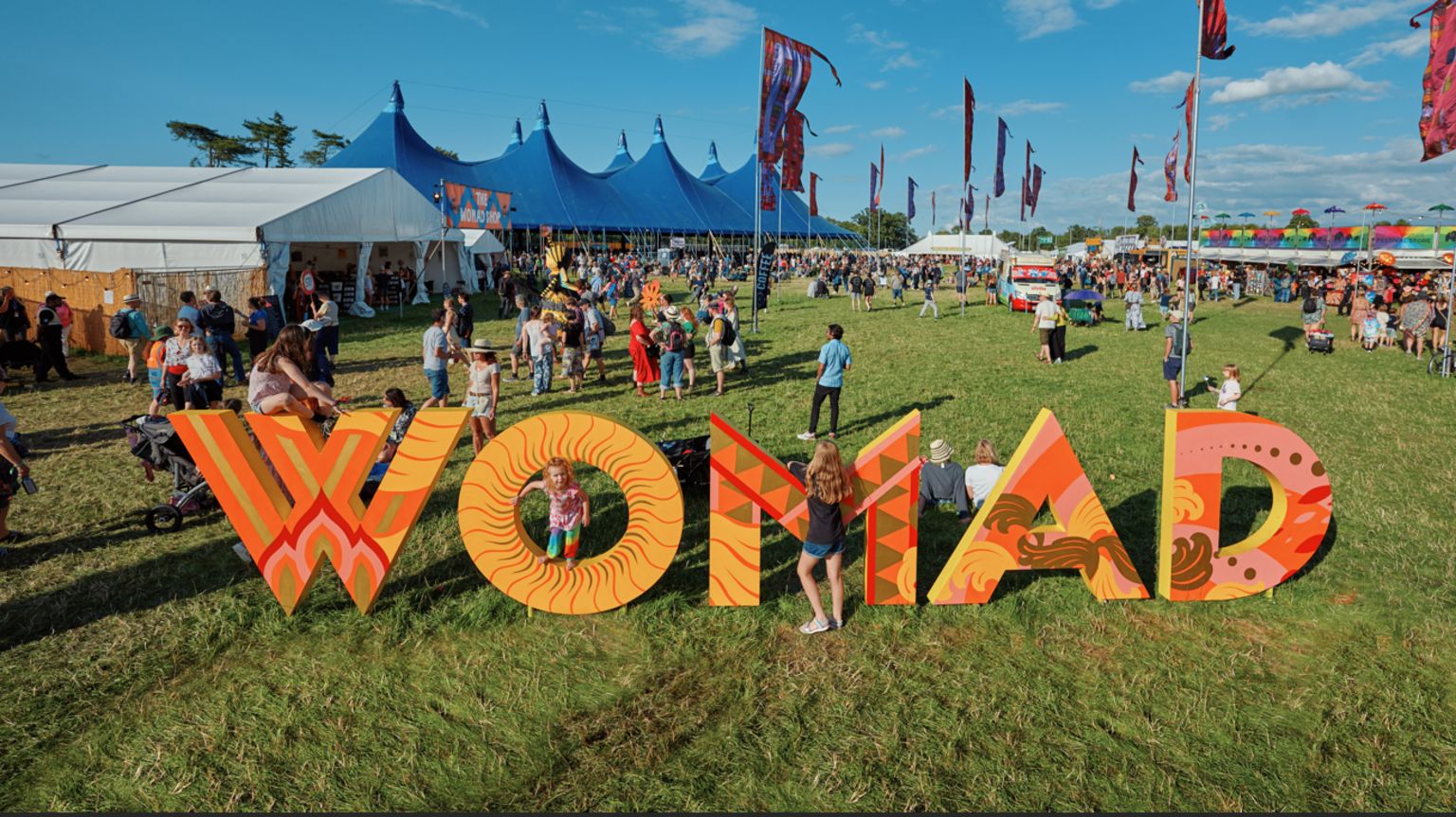 Children playing on a colourful sign that says WOMAD in a festival field