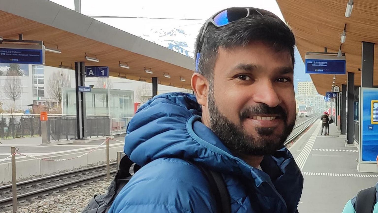 Syed Rafiq, a man with dark hair and a moustache and beard, smiles as he stands at a train station with a blue padded jacket on and sunglasses on the top of his head 