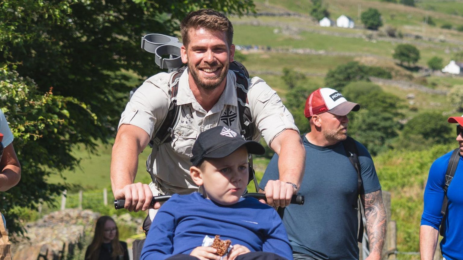 Jack Fleckney with short brown hair and a beard wearing a grey T-shirt and carrying a rucksack. He is pushing a wheelchair in which Albert is sitting, wearing a blue cap and a blue top. Fields on the edge of Snowdon are visible behind them