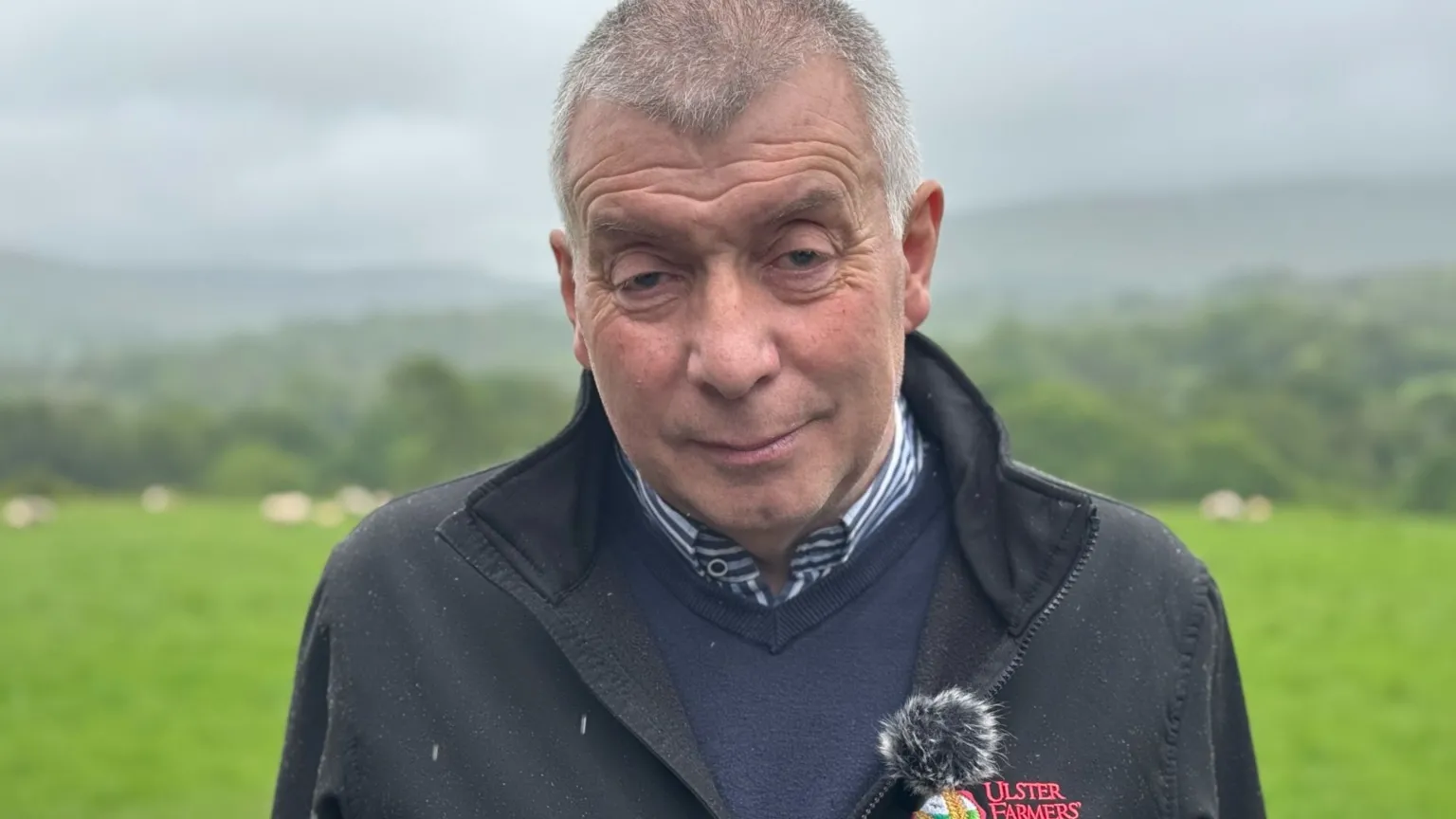 A man with grey hair, he is wearing a black jacket, navy jumper and blue and white shirt