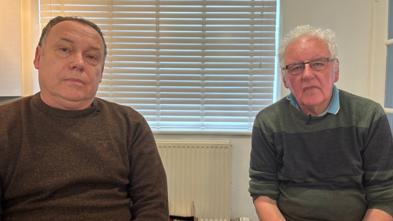 Sion Tecwyn sat on left wearing brown sweater, with Noel on the right in green sweater with blue collared polo shirt, both sat in front of a window with blinds shut at Noel's home at Gaerwen,  Anglesey
