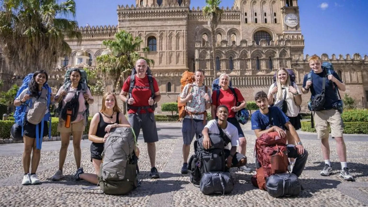 The 10 contestants taking part in the latest series of Race Across the World are gathered for a photograph in front of palm trees and a Gothic-style building. They all have large backpacks and are smiling at the camera.