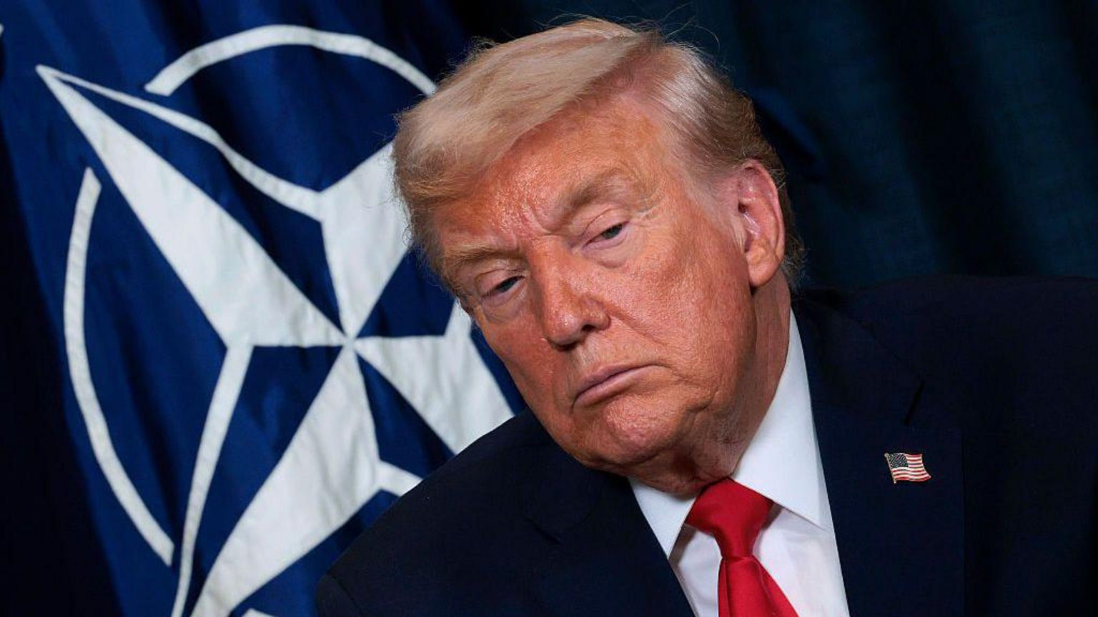 美国总统特朗普身穿深色西装、打着红色领带，背景可见北约旗帜
