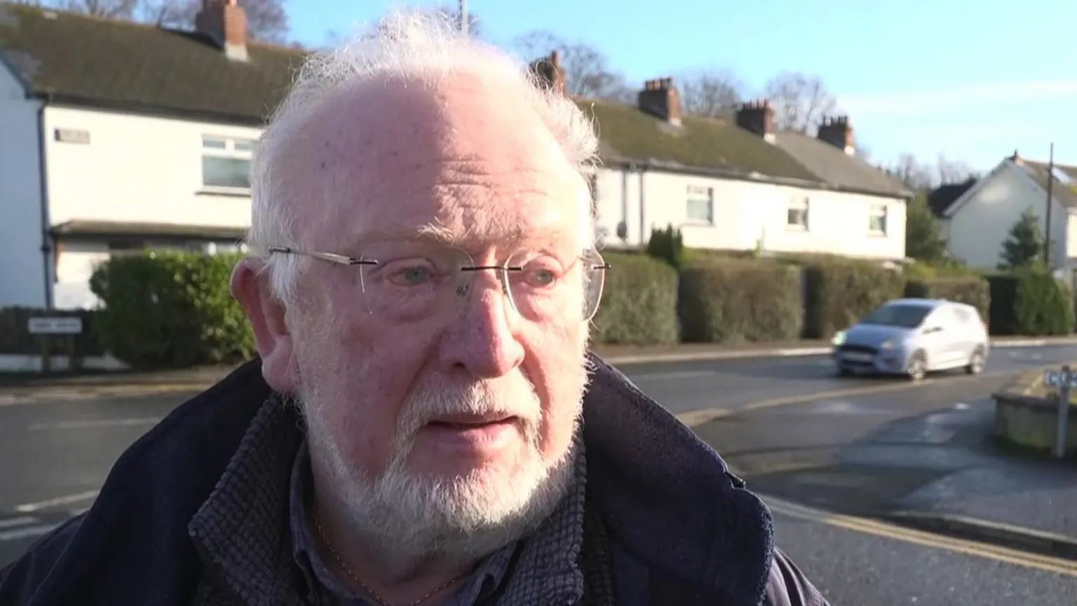 Man with white hair and white beard. He is wearing light framed glasses. He is wearing a purple coat.