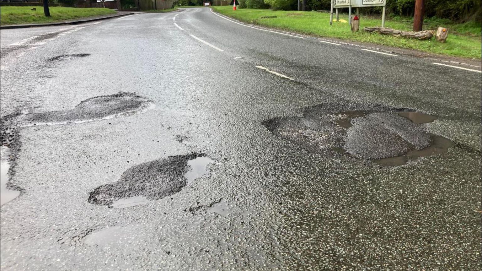About five potholes on a road. The potholes are on both sides of the road and the image also shows grass and the poles of a sign but not the entire sign
