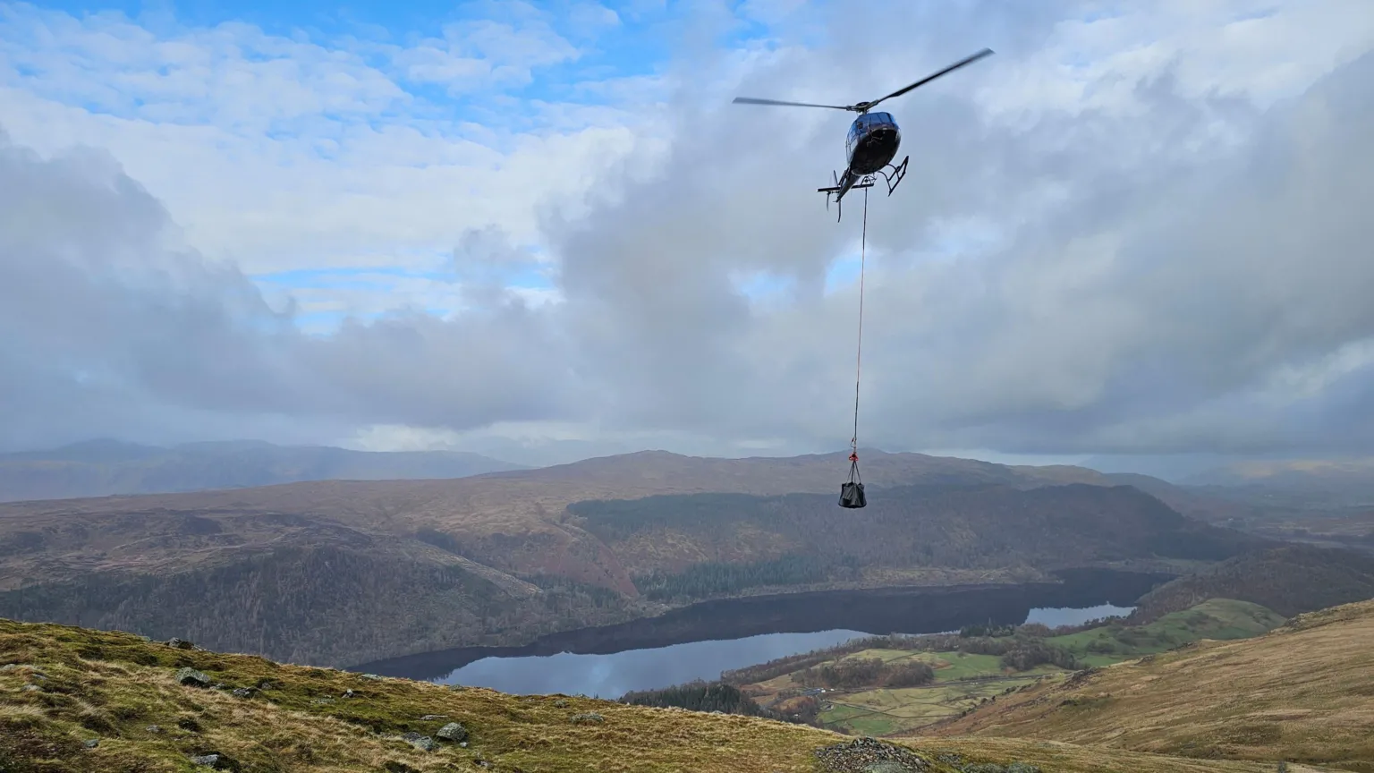Fix the Fells A helicopter flies over Helvellyn. It has a rope hanging down as it transports a quantity of stone. A lake - Thirlmere - can be seen in the background.