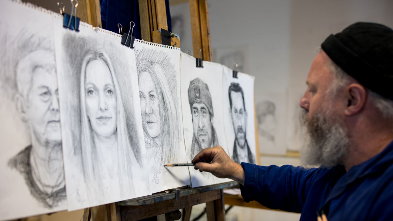 A man drawing a woman's face on an A3 piece of paper. In front of him are four other pieces of paper with people's faces on. 