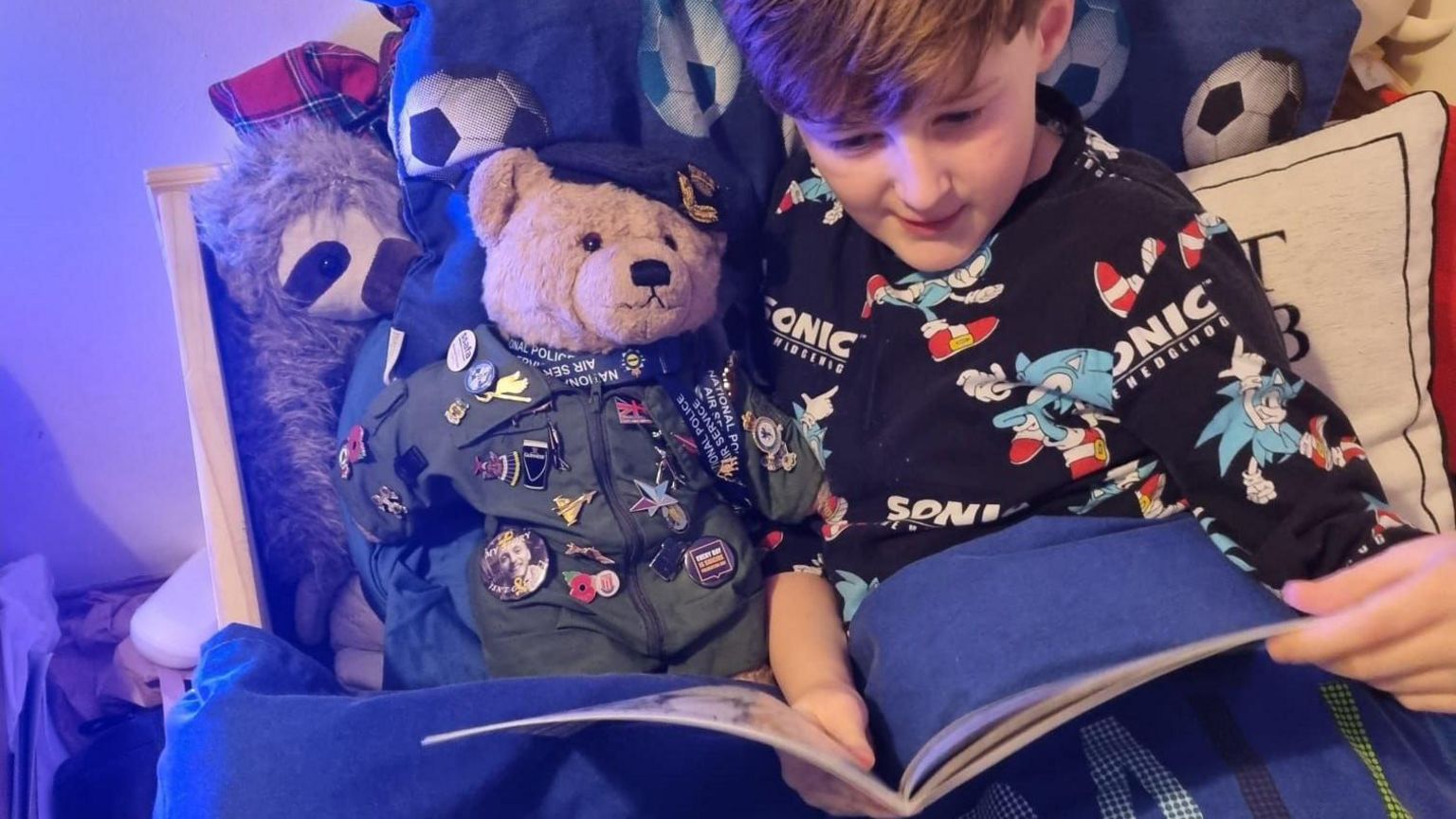 A young boy is reading a book in bed to a mascot teddy bear.