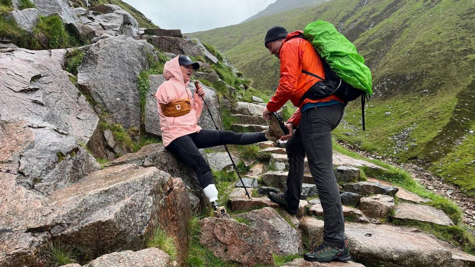 Sarah de Lagarde is helped with her hiking boot while sat on a rock