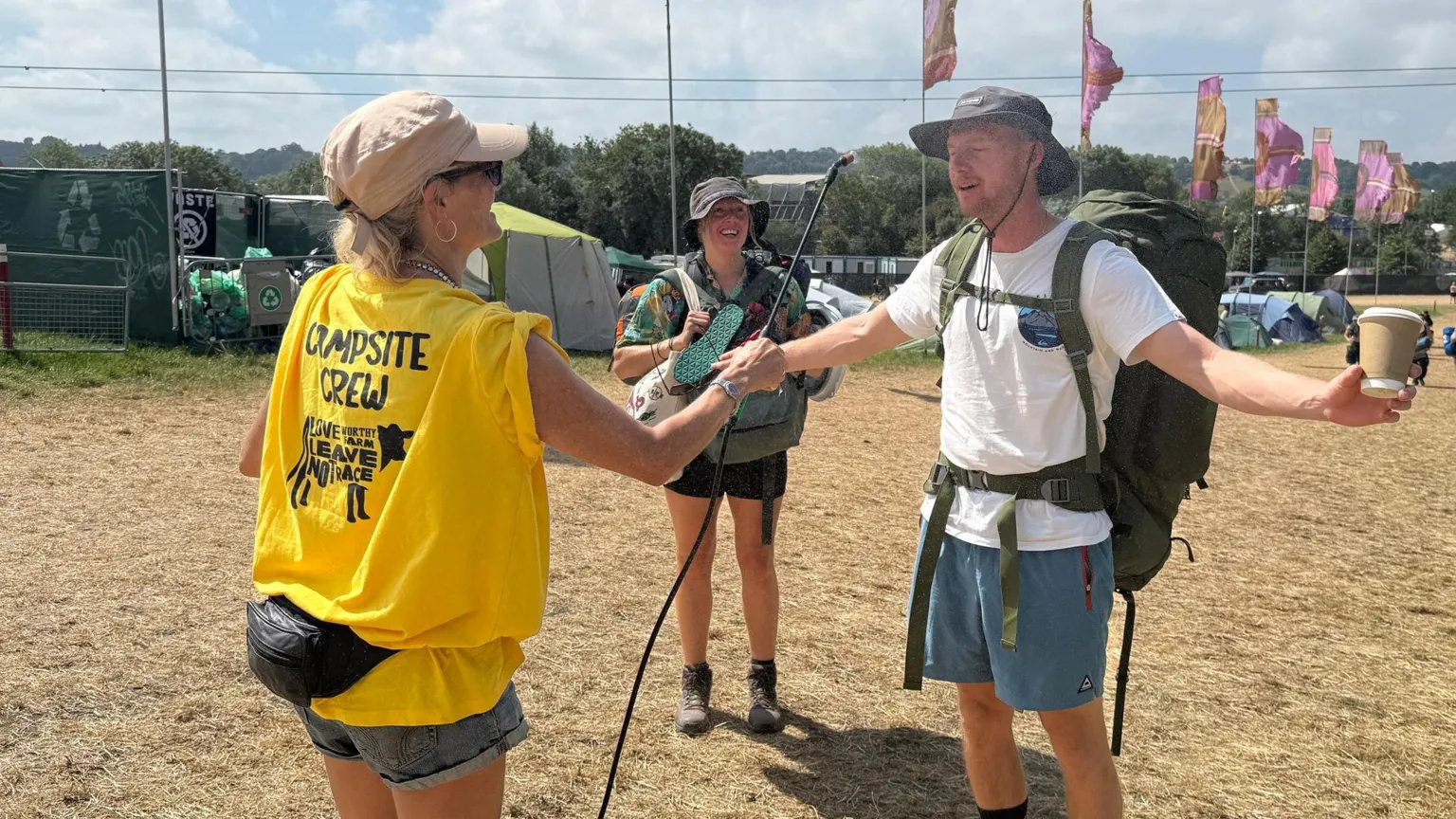 A woman wearing a yellow t-shirt that reads "campsite crew" sprays water over a man's face. The man is wearing blue shorts, a white t shirt, hat and rucksack. He stands with his hands outstretched holding a coffee cup. A woman stands to his right in shorts and carrying bags. In the background is a stretched of browned grass and pink flags.