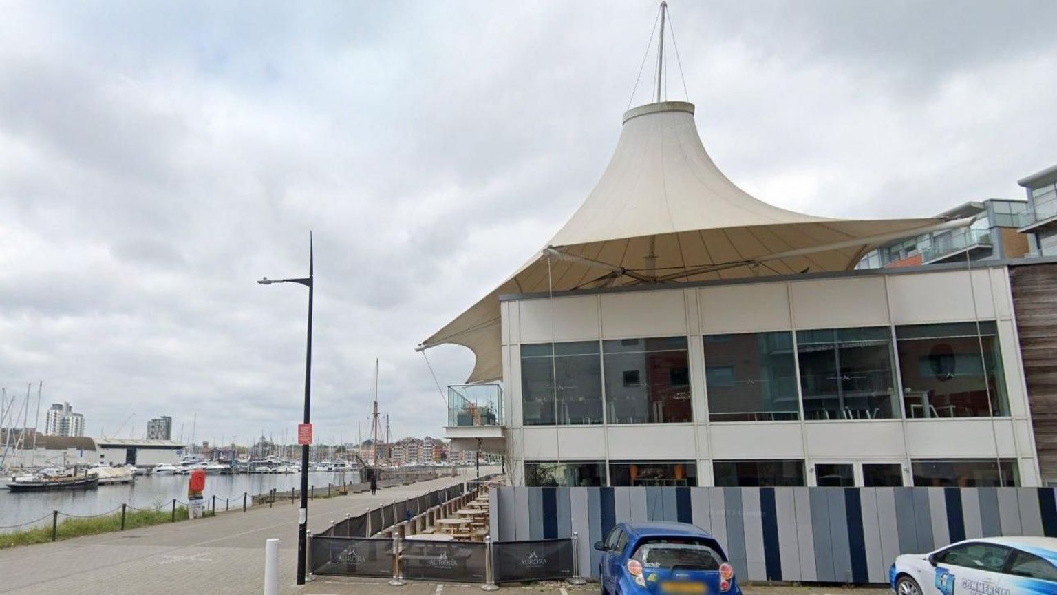 The side of a bar glass-panelled bar with a pergola roof on a waterfront, with two cars parked outside