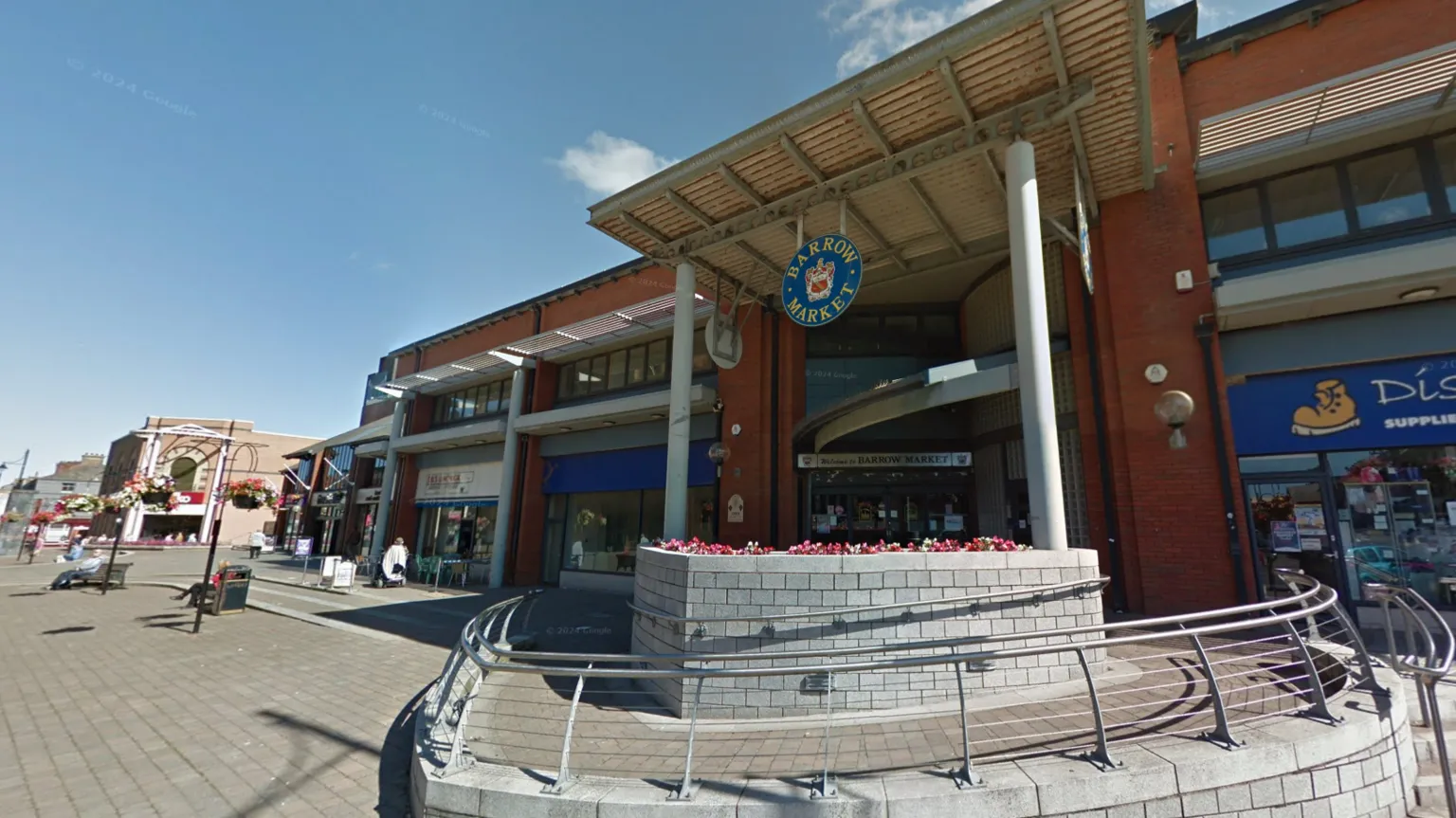 Google Barrow Market Hall pictured from the street outside. There is a grey stone planter containing red flowers in front of the hall, which is a red brick building with metal pillars holding up the roof. In the centre dangles a round blue sign with the words Barrow Market and a heraldic shield. Shop fronts span out from a central entrance.