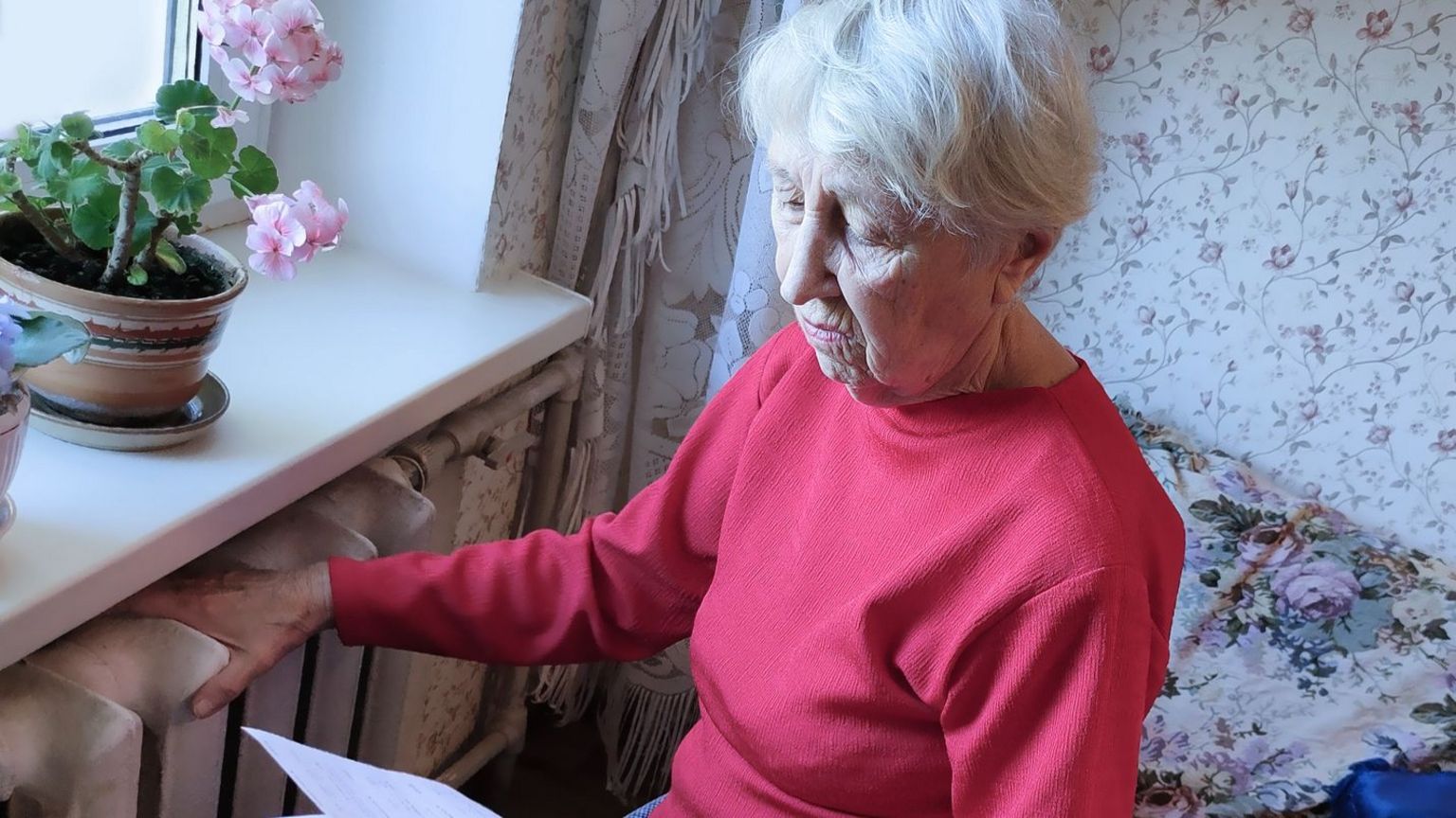 An elderly woman looks at a piece of paper while also feeling the temperature of a radiator