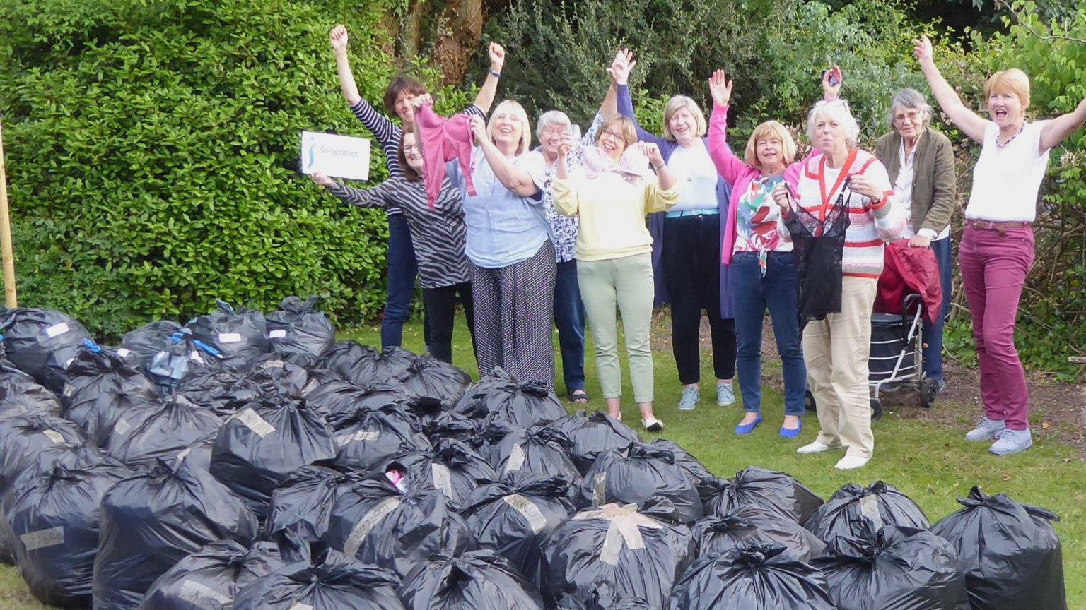 Ten women, some holding bras in the air and others with their arms in the air in a garden standing behind several dozen black bags stuffed with donated pre-loved bras. 