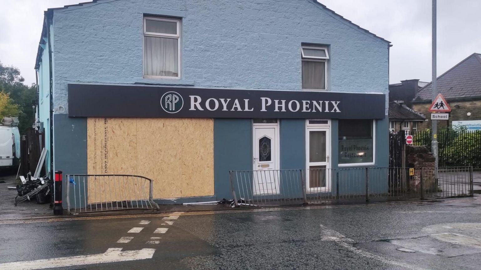 The restaurant's front window has been boarded up. The front of the building is painted blue, with a white door and the sign 'Royal Phoenix' across the top. The railings outside the shop are crumpled after the crash.