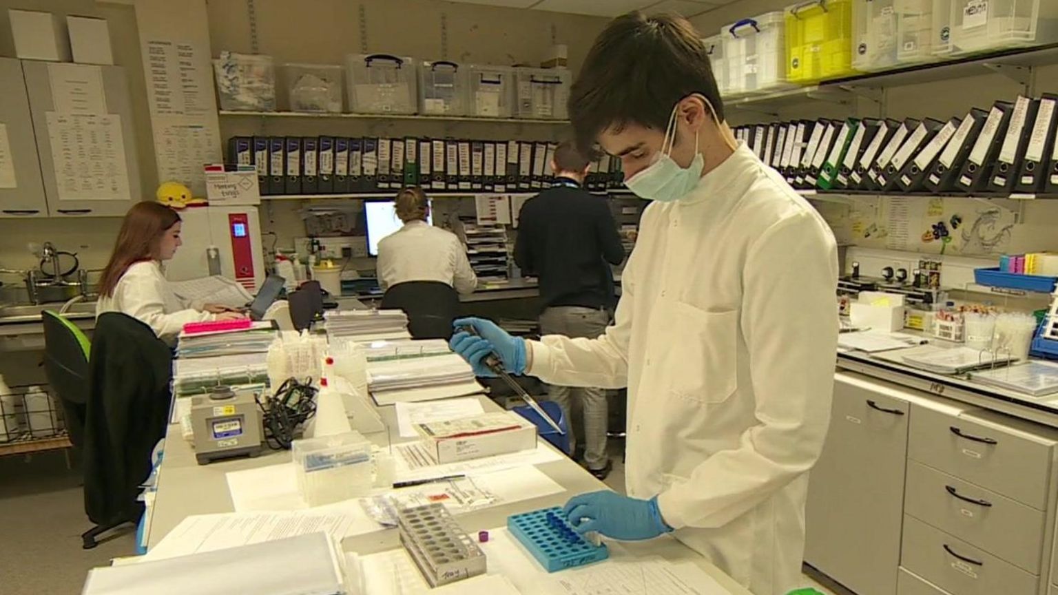 A male scientist wearing a white lab coat and blue gloves is holding a pipette in one hand and a vial in another. In the background are other people sat looking at computer screens. There is a shelf filled with lots of folders on the wall.