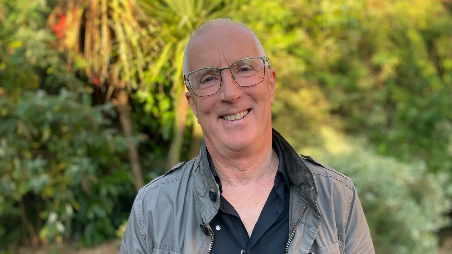 A man is smiling. He is wearing glasses, a grey jacket and black shirt. He is standing in front of trees and greenery.