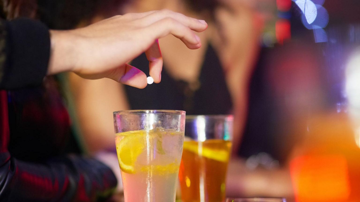 A hand holding a small white tablet over a glass of drink at a bar.