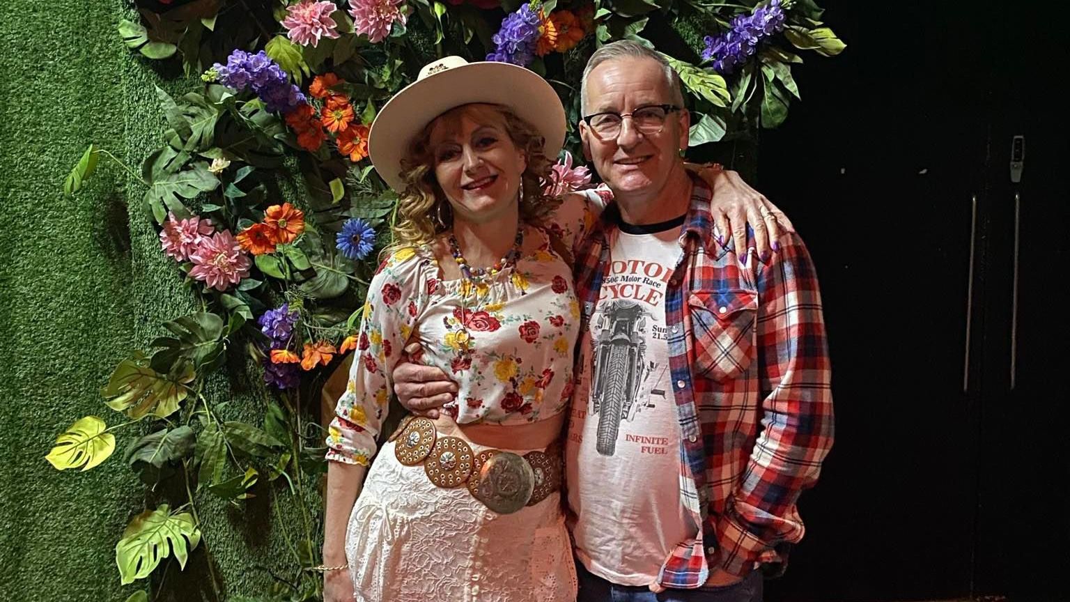 A woman in a hat and floral blouse, with arms around a man in a checked red and blue shirt and motorcycle t-shirt