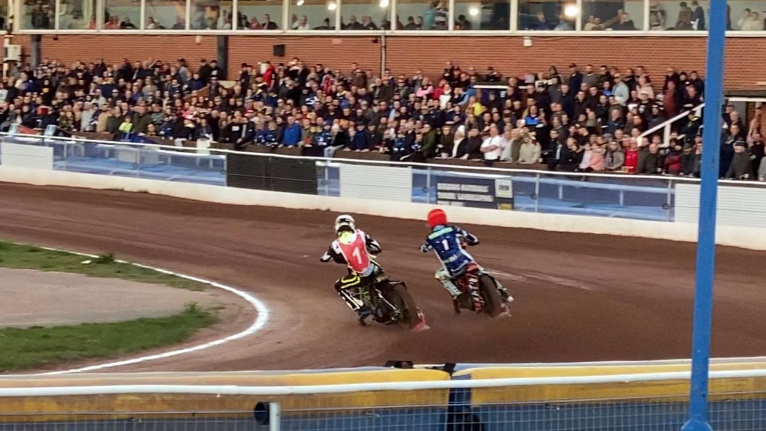 Two speedway riders drifting on the bend while spectators watch from stands