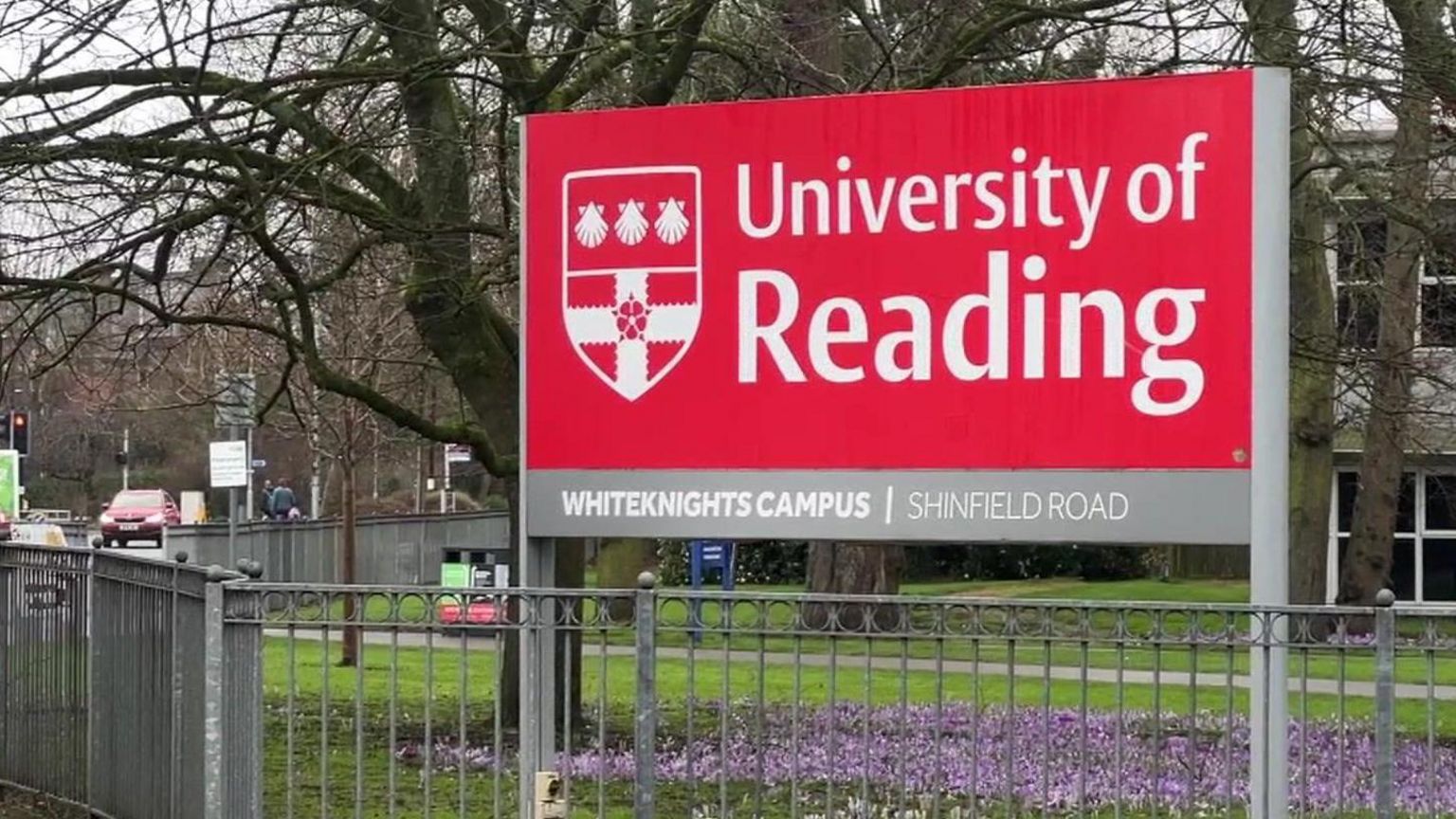 Red sign which reads University of Reading with a shield insignia. The sign is by a road. In front of the sign there is a metal railing. There are purple flowers underneath.