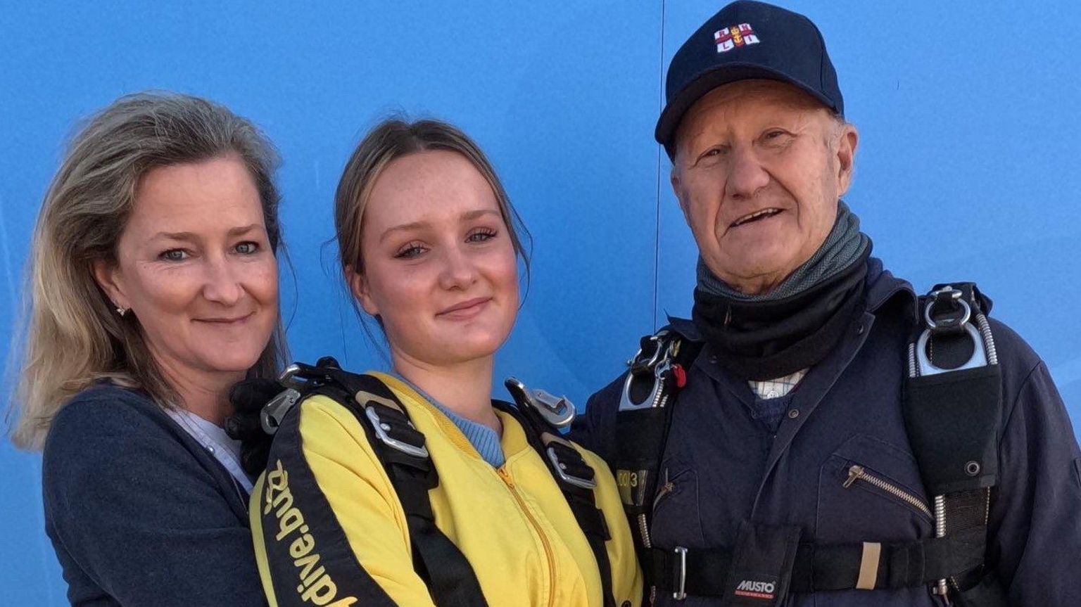 Mr Copeland, his daughter Melanie Exon, and granddaughter Annabel Exon standing against a blue background. Annabel and Mr Copeland are both wearing skydiving suits and Mr Copeland has an RNLI hat on