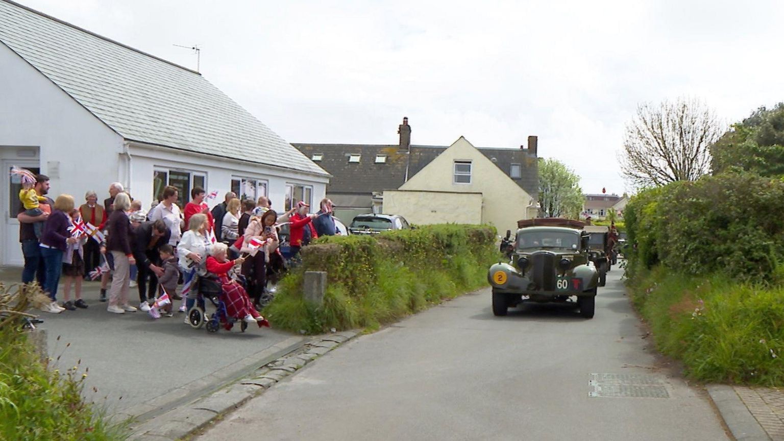 Old vehicles driving down a road while people watch from a driveway