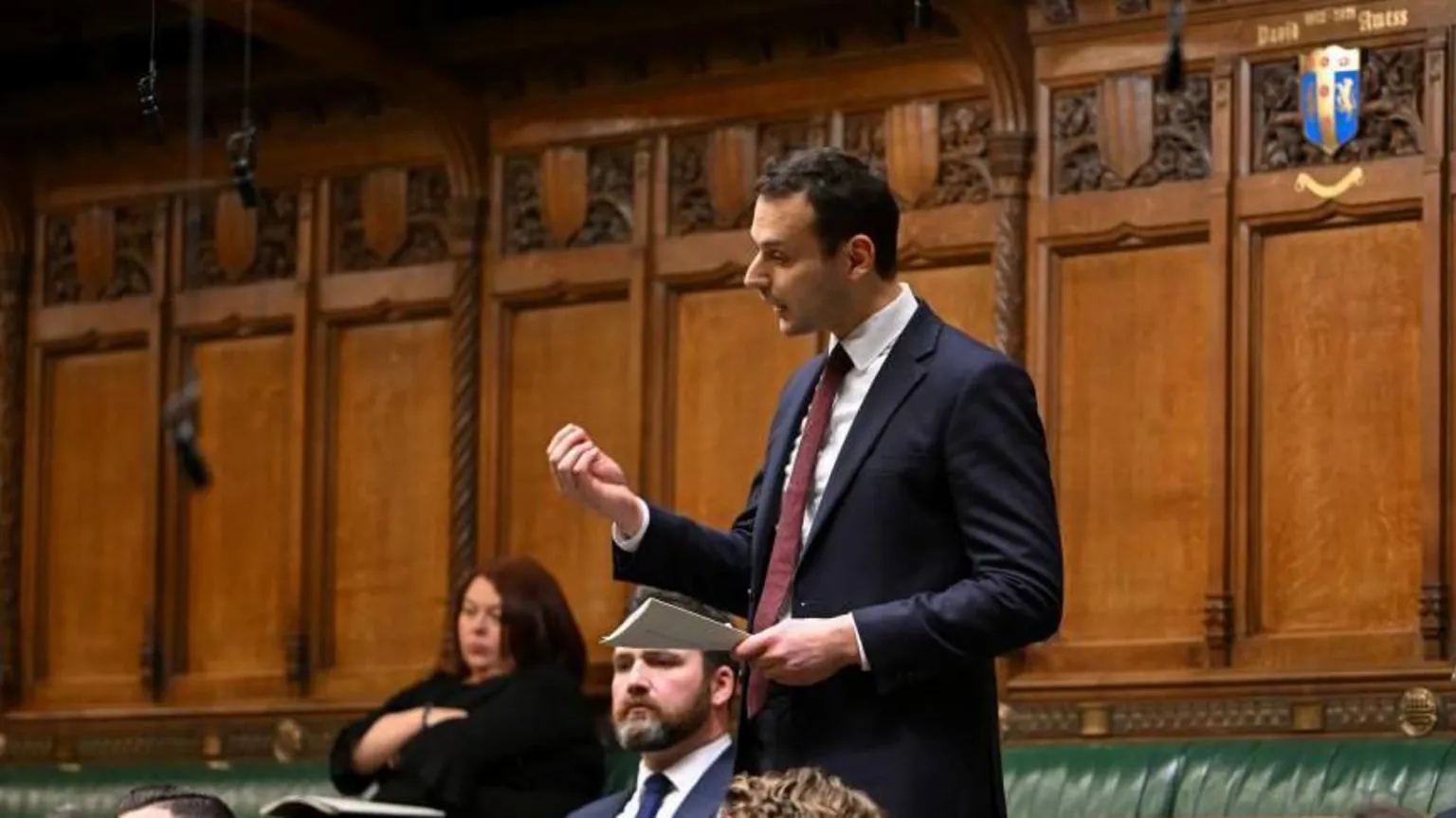 Reuters Labour MP Josh Simons speaks in the House of Commons. He is wearing a dark blue suit, burgundy tie and white shirt. He holds a piece of paper in his hand and gesticulates with his hand as he speaks. Other MPs sit on the green benches around him.