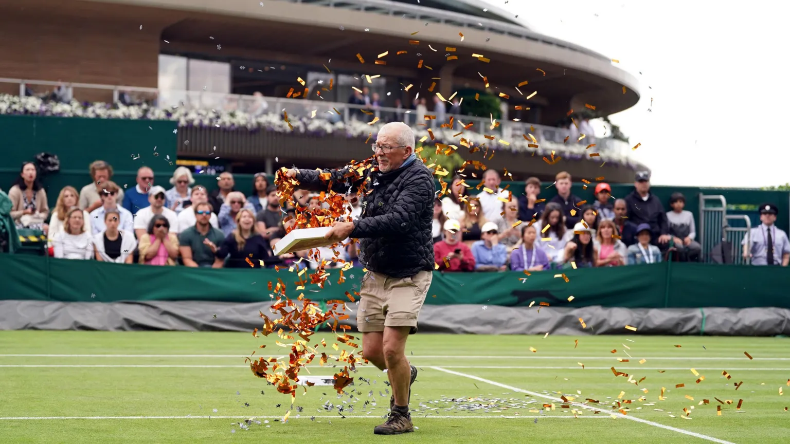 An elderly man in dark jacket and beige shorts runs across a grass tennis court, scattering orange and yellow confetti from a container. Spectators watch from green-bannered stands under a curved roof structure. The confetti creates a colorful trail across the pristine court surface.