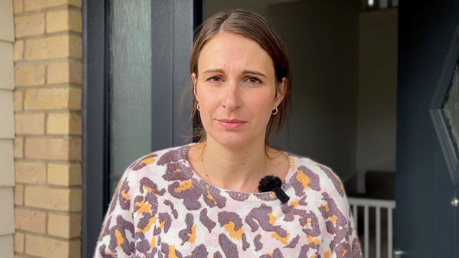 A woman, Kelly Heather, wearing a colourful top and standing in the doorway of her home