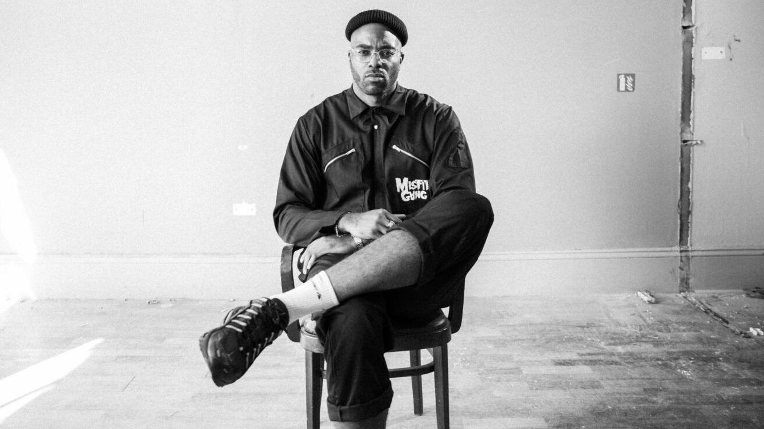 Moody, black and white image of Chiedu Oraka sitting, with legs crossed, on a chair in an otherwise empty room