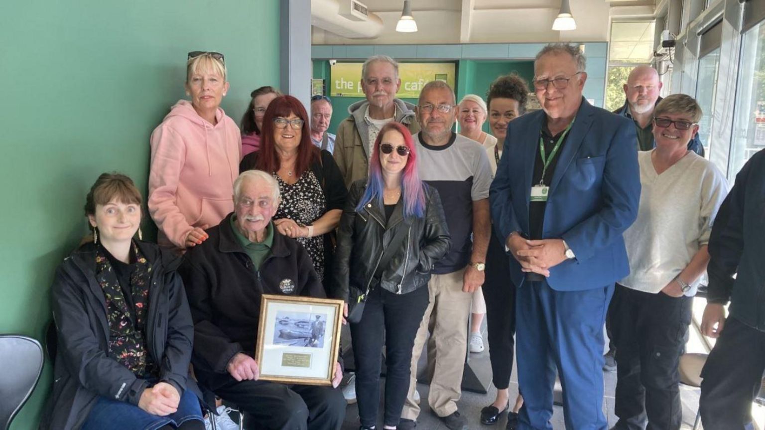 Stuart MacDonald smiling with his frame with an old photo of him maintaining the park, surrounded by several colleagues in The Pavilion Cafe at East Park