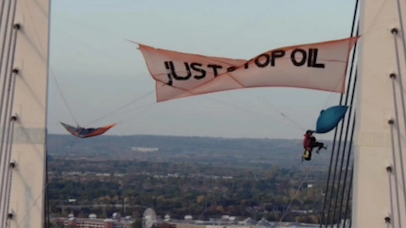 The image shows protesters on a high bridge or tower structure with a large banner reading 