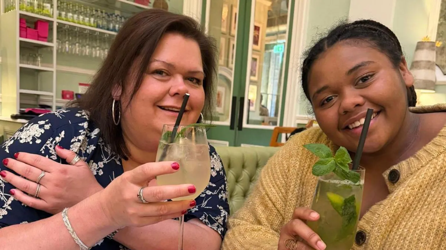 Family photograph Caroline with her daughter Francesca. They are both holding Mojito cocktails. One is non-alcoholic. Caroline has a navy blue flowery dress and silver hooped earrings and Francesca has a fluffy yellow cardigan. They are in a bar.