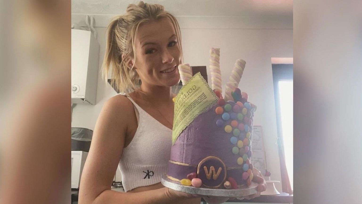 A young blonde woman with her hair tied in a ponytail. She is smiling at the camera and holding a cake covered in Marshmallows, chocolate sweets and a piece of yellow paper which reads "Golden Ticket".