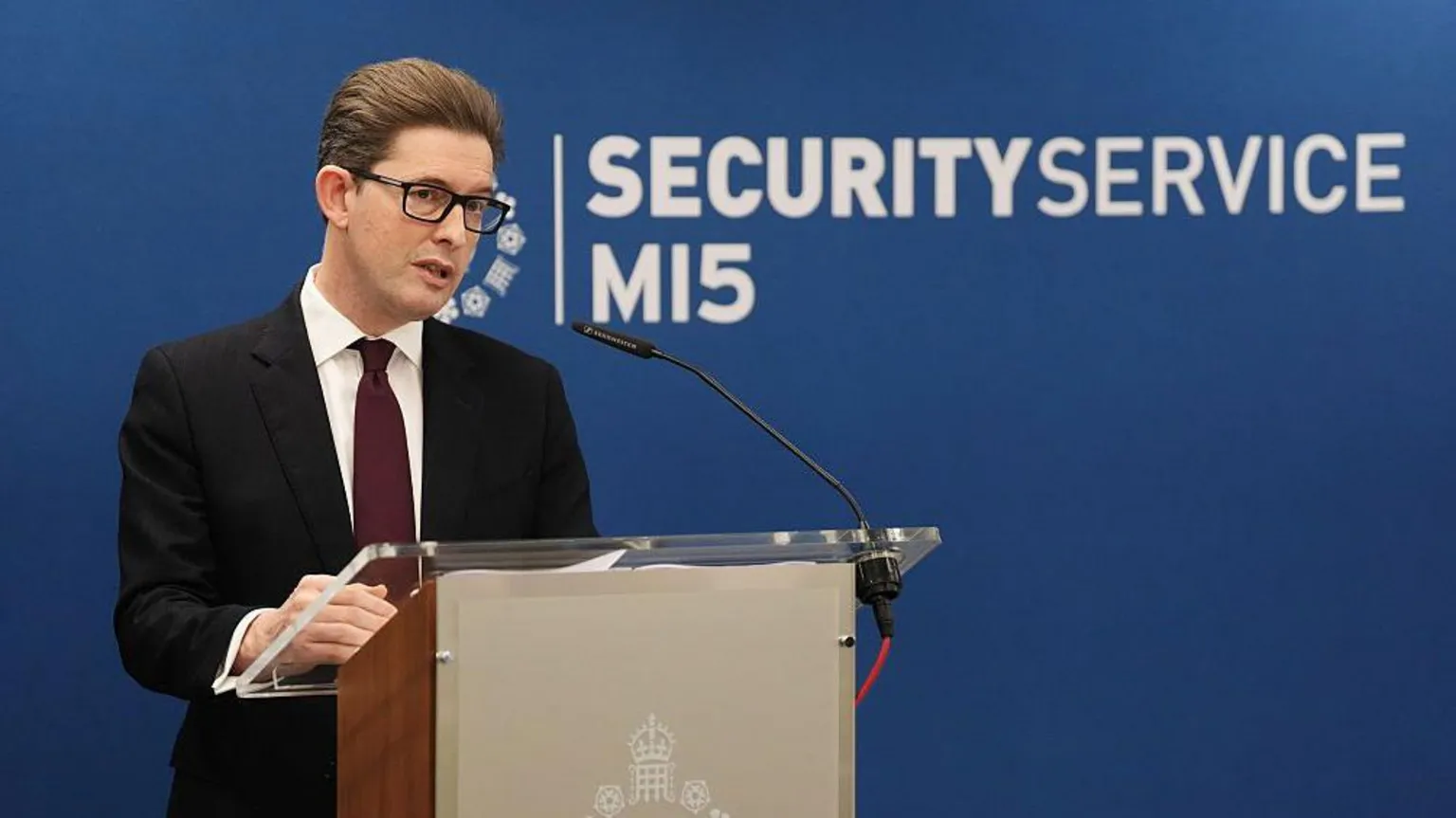 Getty Images Sir Ken McCallum standing behind a podium with the MI5 insignia emblazoned on the front in front of blue background with the words 'MI5 security service' behind him. Sit Ken is wearing a black suit, a dark maroon tie, white shirt, black-framed glasses and clicked back brown hair.