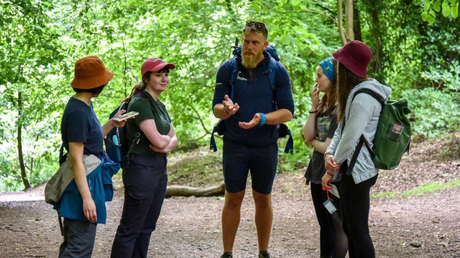 A group of walkers standing together in a clearing in woodland. Some are wearing backpacks, hats and waterproof jackets tied about their waist